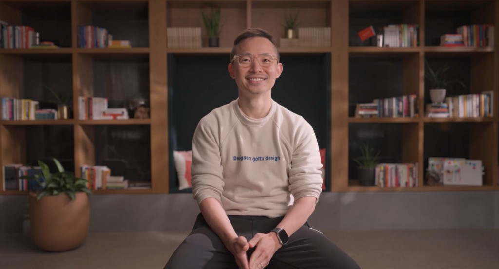 Weinan sits and smiles in front of a bookshelf, wearing a sweatshirt that reads Designers gotta design.